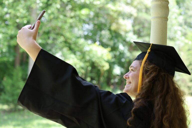 Graduate in cap and gown taking a selfie with a smartphone outdoors, smiling in a sunny, leafy setting