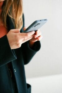 Close-up of a person holding a gray smartphone with both hands, wearing a dark coat, light blurred background.
