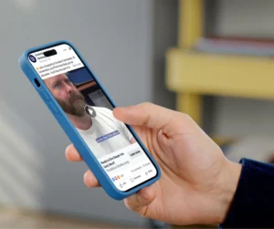 Close-up of a hand holding a blue smartphone displaying a bearded man in a video post on a social feed, with like/comment icons visible.